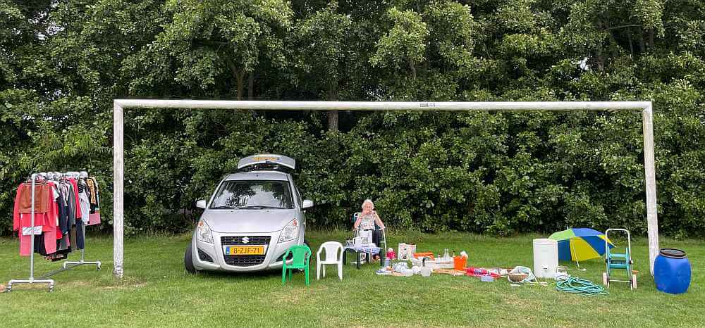 Oude vrouw zit in Hoorn op Terschelling op een rommelmarkt tussen haar spullen zonder publiek.